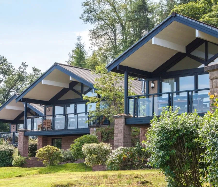 Exterior of the Cameron Lodges overlooking Loch Lomond, with lush green lawns and bushes in the foreground.