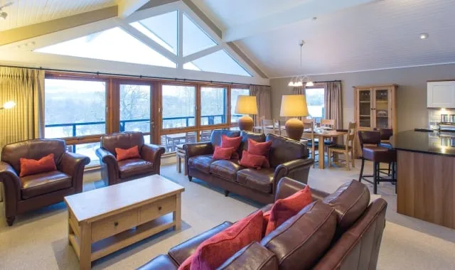 Interior of the three bedroom lodge, featuring brown leather sofas with red pillows around light brown table. A balcony overlooking Loch Lomond is visible through the glass French door.