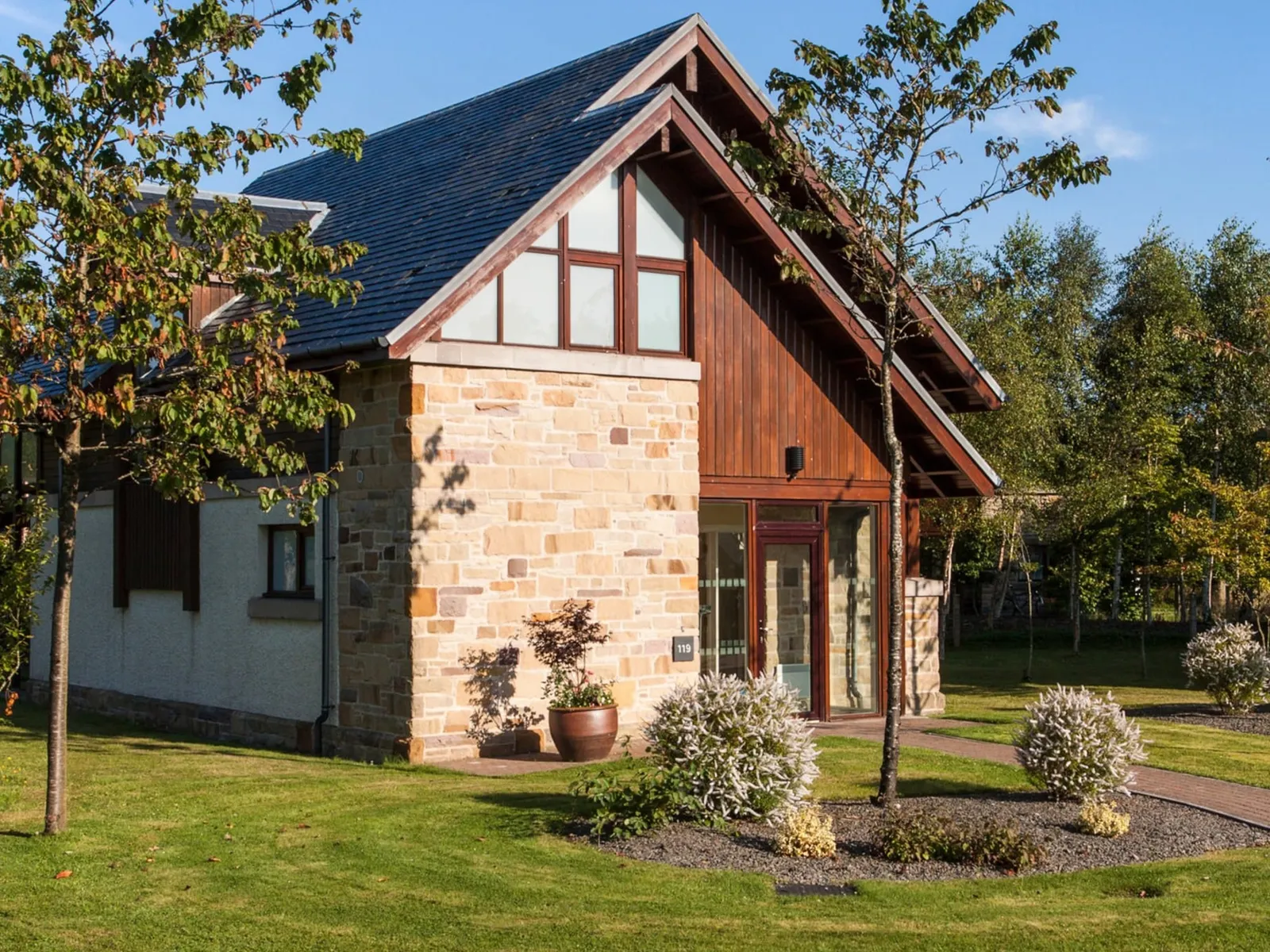 A modern, luxury detached self-catering Loch Lomond lodge with Scottish architectural features. The two-story exterior combines warm wood paneling and rustic golden stone cladding under a steep slate roof. The lodge is set within a well-manicured green lawn and surrounded by mature trees, suggesting a tranquil holiday setting on the shores of Loch Lomond.