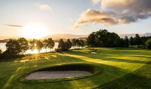 A stunning view of the Carrick Golf Course at Loch Lomond at sunset. The meticulously maintained green fairway is visible in the foreground, featuring a large, oval-shaped sand bunker. The bright sun is setting low over the water of the loch on the left, casting long shadows across the grass.