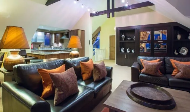 The expansive, open-plan living and dining area of a luxurious three-bedroom detached lodge at Loch Lomond Shores. The room features dark leather sofas adorned with orange and paisley-patterned cushions, set around a wooden coffee table. Two large lamps with wide, amber shades provide warm lighting. The modern kitchen with wood cabinetry is visible in the background, along with built-in dark wood shelving and exposed ceiling beams.