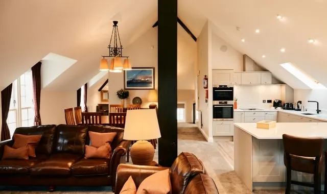 The bright and airy open-plan living space of the Three Bedroom Coach House at Cameron Club Lodges at Loch Lomond. The foreground features a rich brown leather sofa with orange cushions. The vaulted, white ceiling highlights an elegant chandelier above the wooden dining table in the mid-ground. To the right, the modern, integrated kitchen is visible with light grey cabinetry and a white central island, all separated by a dramatic, central dark support column.