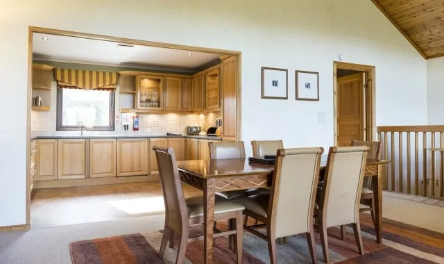 Well lit dining area of two bedroom detached lodge, featuring a dining table with six chairs. Nature surrounding Loch Lomond is visible through the kitchen window.