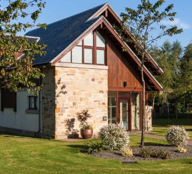 A modern, luxury detached self-catering Loch Lomond lodge with Scottish architectural features. The two-story exterior combines warm wood paneling and rustic golden stone cladding under a steep slate roof. The lodge is set within a well-manicured green lawn and surrounded by mature trees, suggesting a tranquil holiday setting on the shores of Loch Lomond.