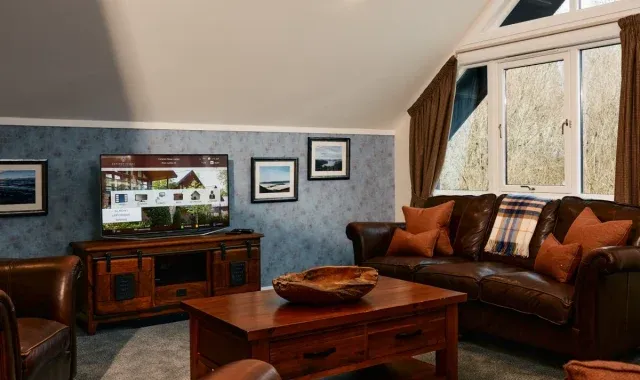 The cosy and luxurious interior of a four-bedroom lodge at Cameron Club near Loch Lomond. The living room features a dark brown leather sofa and matching armchairs, a wooden coffee table, and a matching media console holding a large flat-screen television. The room has vaulted ceilings, large windows letting in natural light, and is decorated with blue textured wallpaper, framed artwork, and rustic orange and plaid cushions and throws.